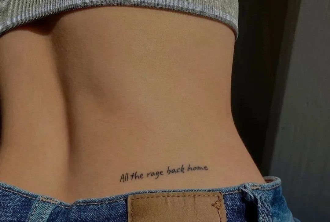 11 Lower Back Script Tattoos for Bold Ink Inspiration 4 A fine-line cursive script tattoo reading 'All the rage back home' on a person's lower back above denim jeans.