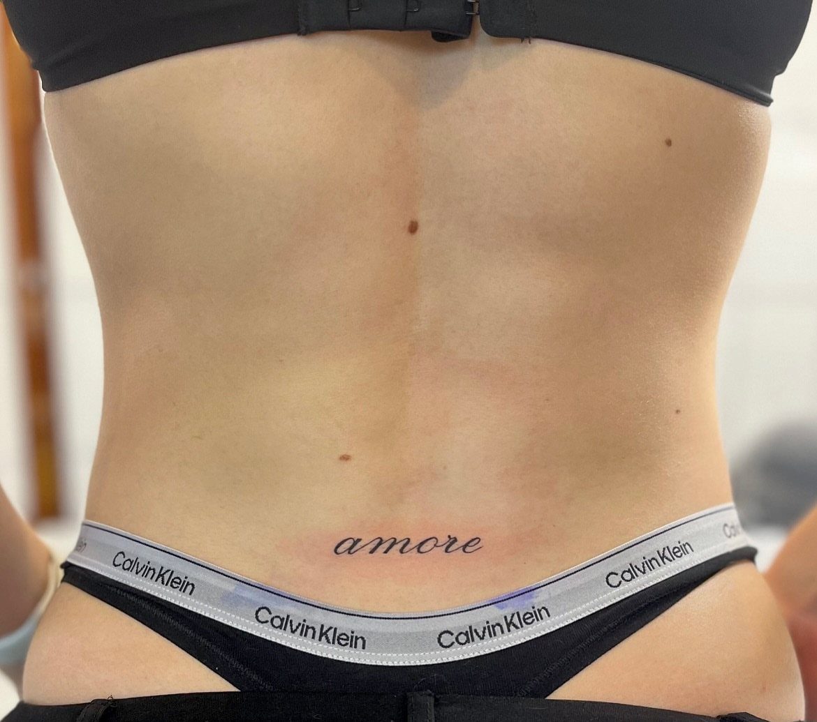 11 Lower Back Script Tattoos for Bold Ink Inspiration 8 A small cursive script tattoo reading 'amore' on a person's lower back.