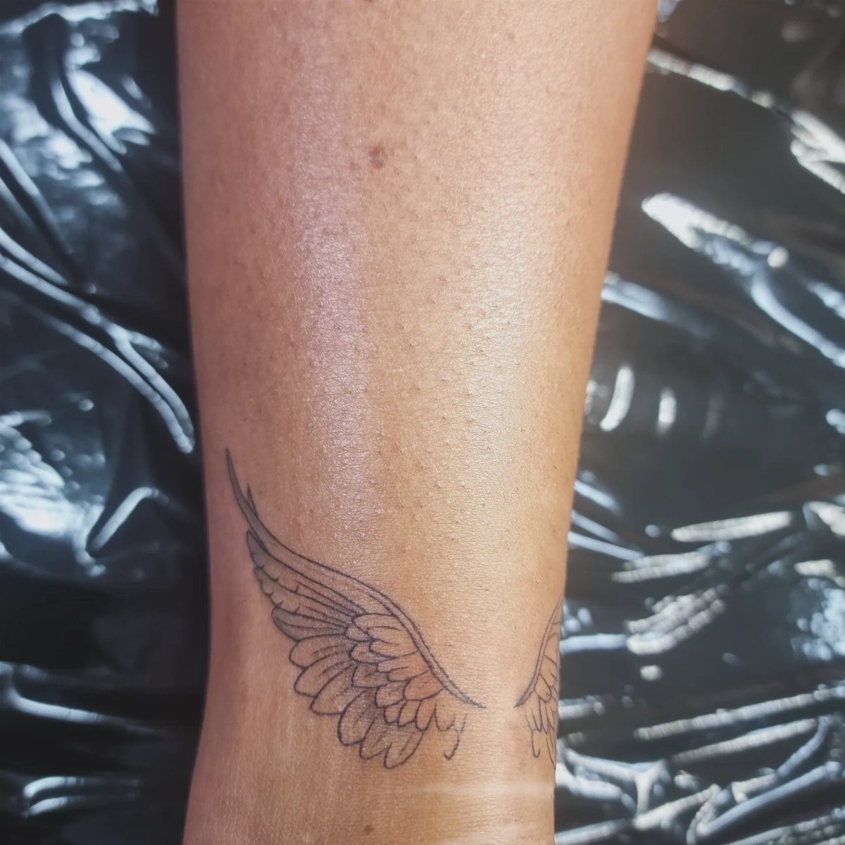 16 Front Ankle Tattoo Ideas for Women 2026 7 A black ink angel wings tattoo wrapped around a person's ankle.