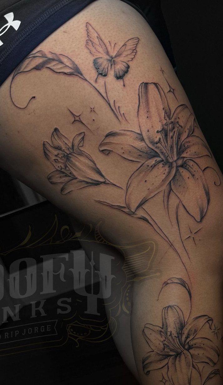 Side Thigh Tattoo: 7 Black Ink Designs That Use the Space 3 Black ink fine line tattoo of lilies and a butterfly on a person's thigh.