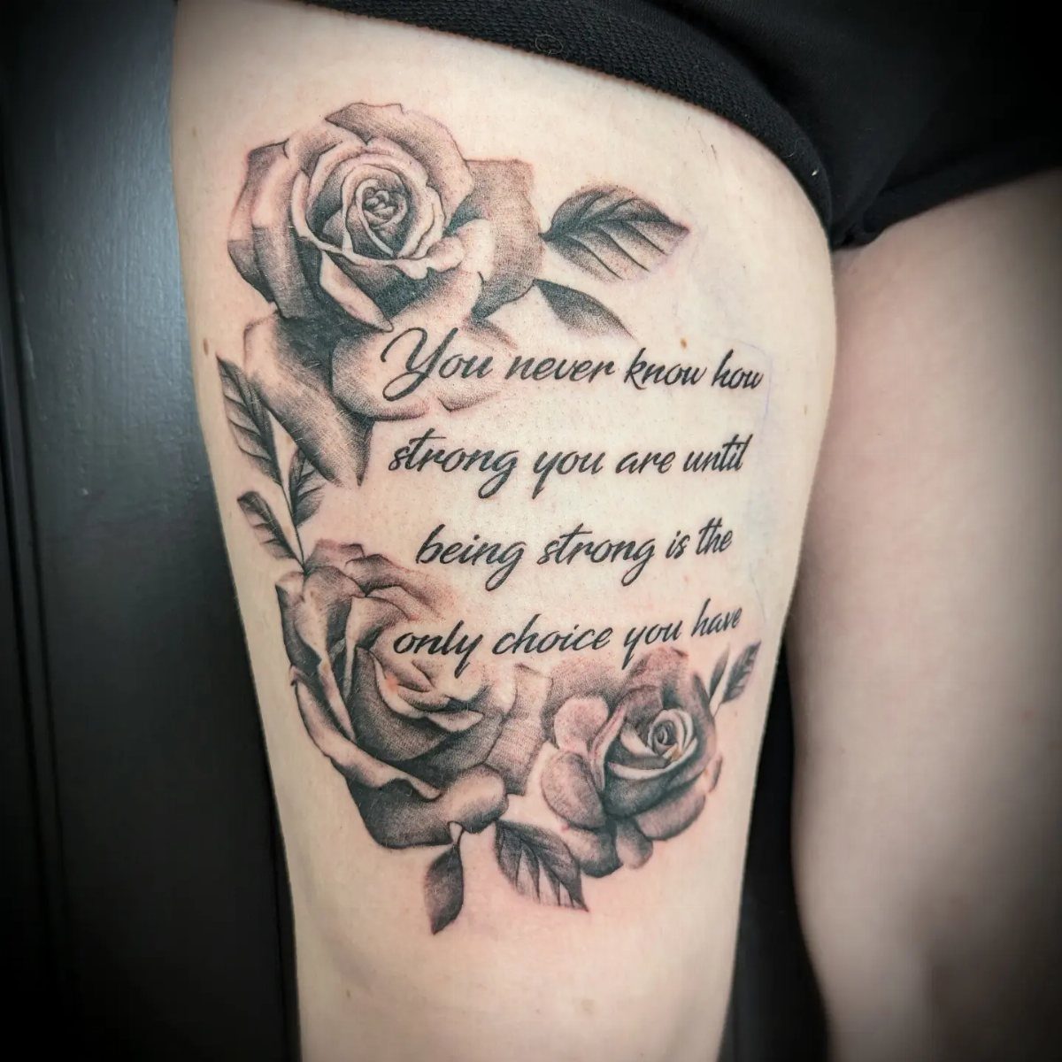 Side Thigh Tattoo: 7 Black Ink Designs That Use the Space 6 Black ink rose tattoo with a motivational quote on a person's thigh.