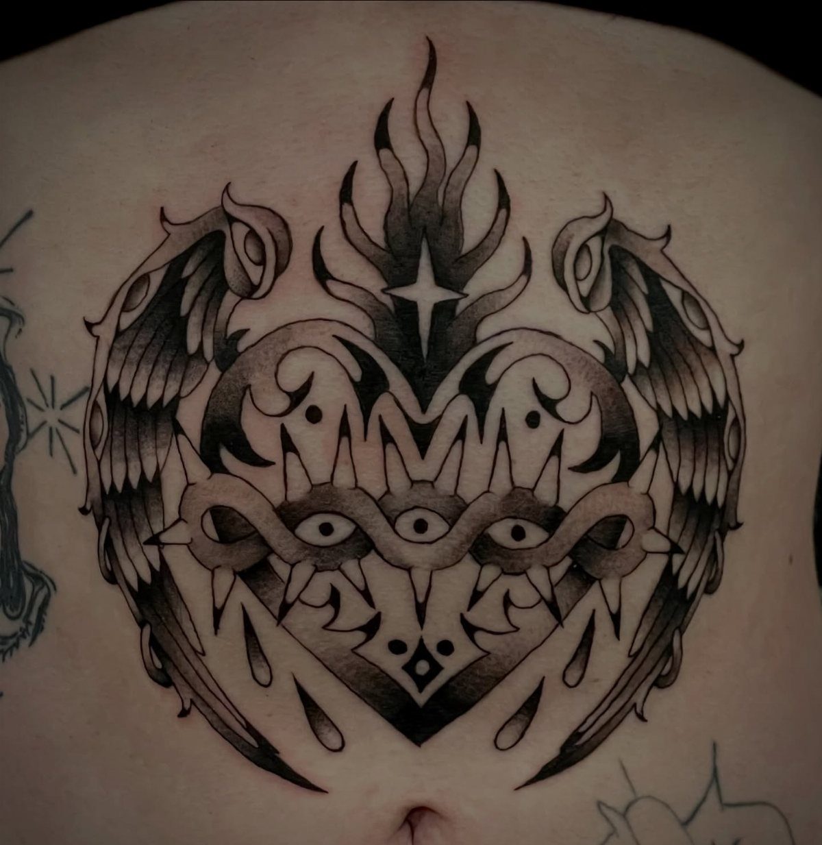 A large symmetrical black and grey winged heart tattoo centered on a person's stomach.