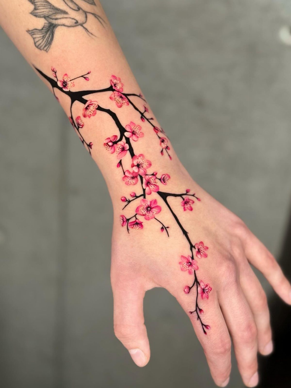 Japanese Tattoo Style: 14 Iconic Motifs and Where They Go 4 A person's arm and hand featuring a detailed pink cherry blossom branch tattoo.