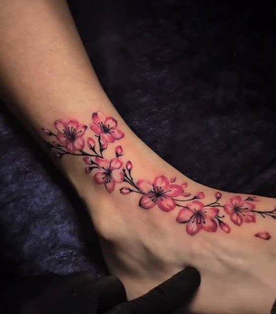 16 Front Ankle Tattoo Ideas for Women 2026 6 Pink cherry blossom tattoo with dark branches winding around a person's ankle and foot.
