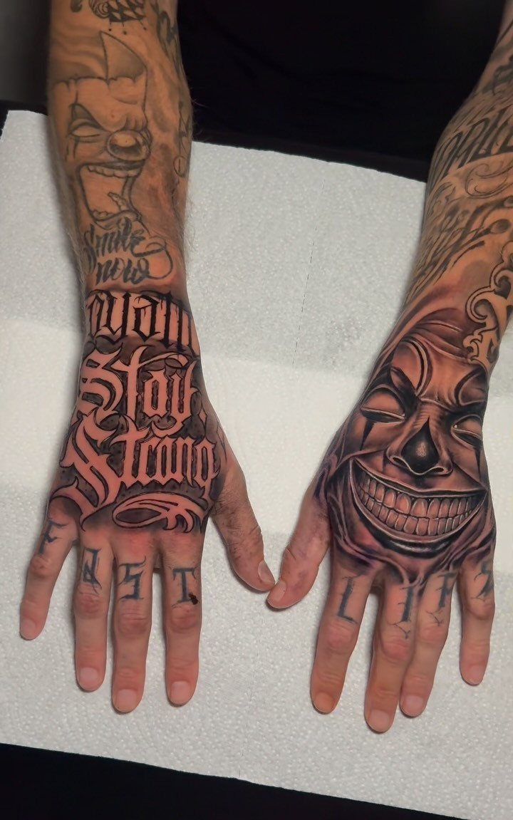 Tattoo Style for Hand: 8 Designs That Work on This Spot 8 Two tattooed hands displaying large black and grey ink designs including lettering and a clown mask.