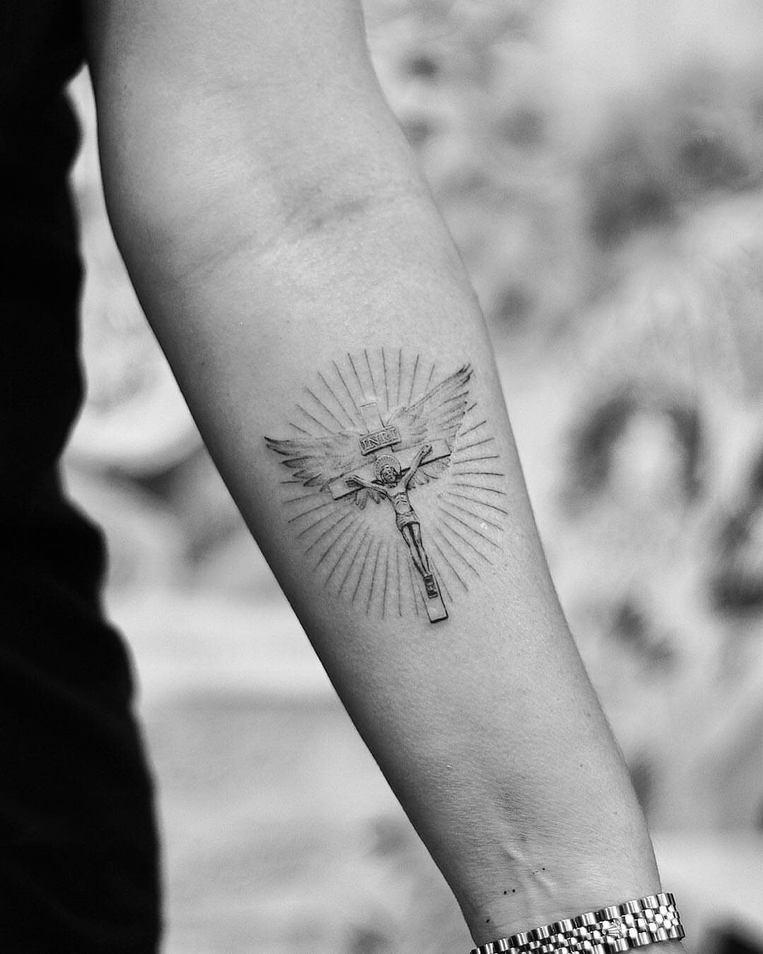 11 Tattoo Arm Women Will Want to See Before Booking 2026 5 A black and white tattoo of a crucifix with wings and sunburst rays on a person's forearm.