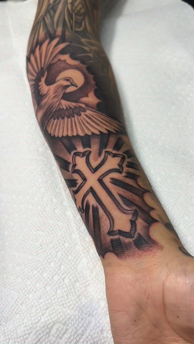 High Contrast Tattoo Design: 8 Bold Ideas That Read From Any Distance 4 A black and grey forearm tattoo featuring a decorative cross and a bird in flight.