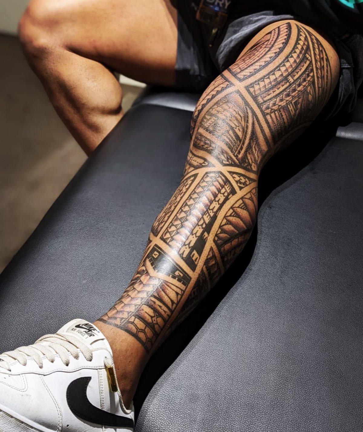 A person's leg covered in a detailed black and grey Polynesian style full sleeve tattoo.