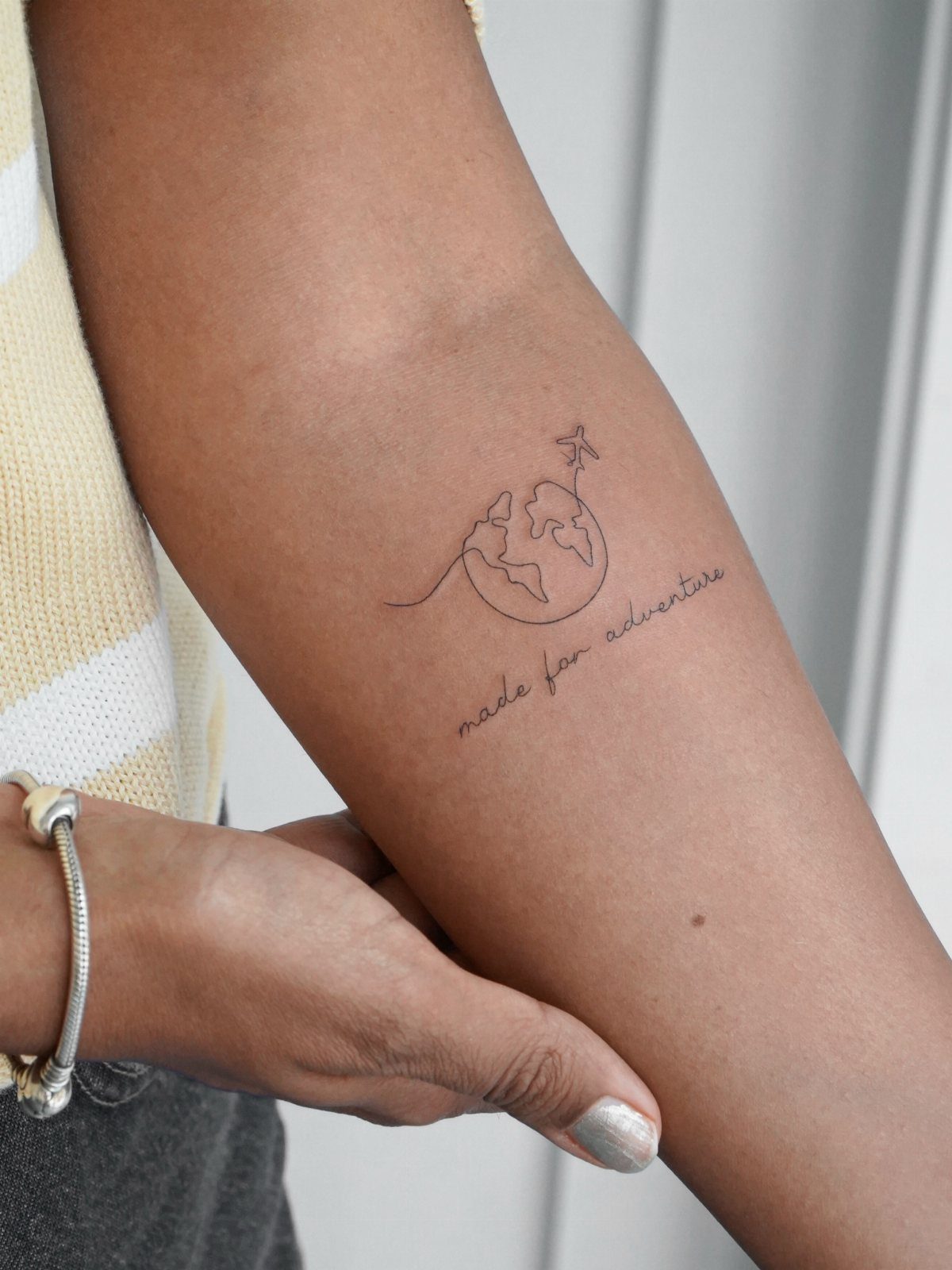 A minimalist fine-line tattoo of a globe and airplane on a person's forearm.