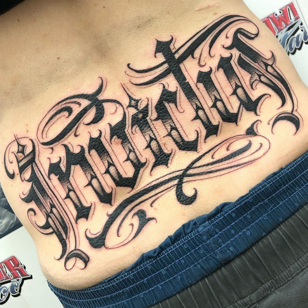 11 Lower Back Script Tattoos for Bold Ink Inspiration 6 Large black gothic script tattoo on a person's lower back above blue waistband.