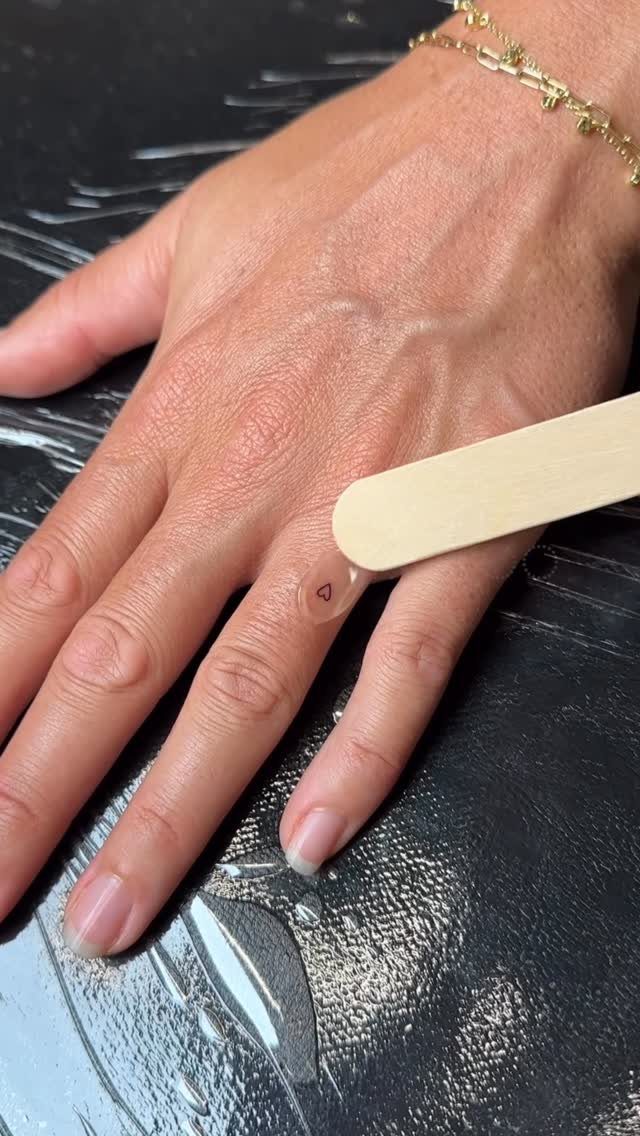 A small, fine-line heart tattoo on the side of a person's ring finger.