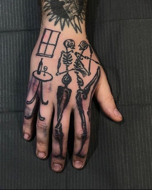 Tattoo Style for Hand: 8 Designs That Work on This Spot 5 A hand featuring black ink ignorant style tattoos of two skeletons at a table.