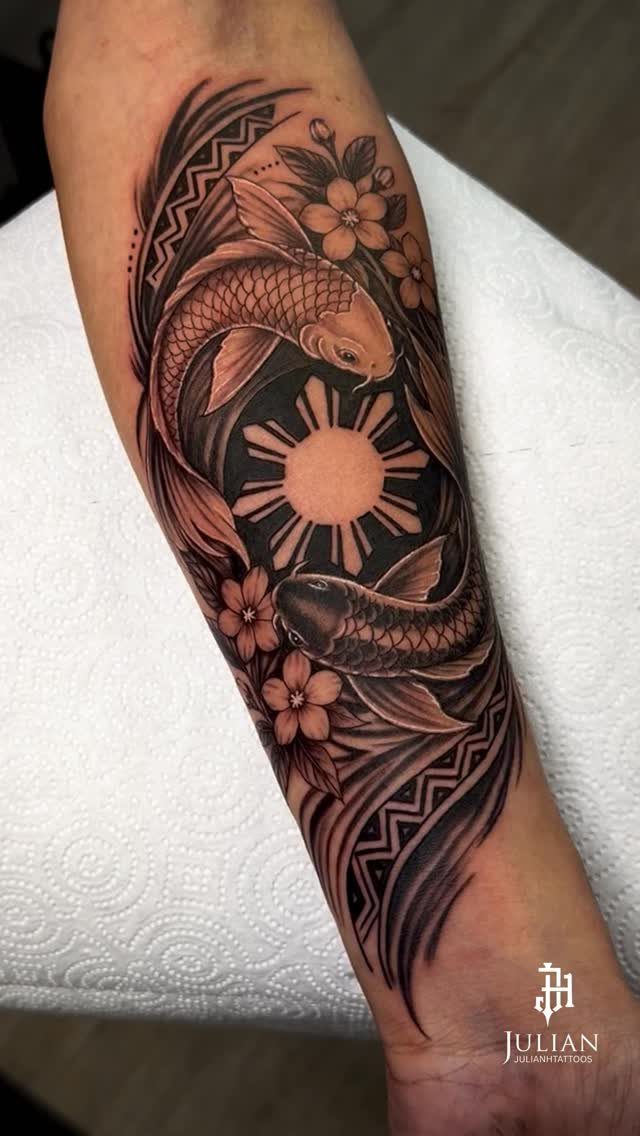 A black and grey forearm tattoo depicting two koi fish circling a sun and floral elements.
