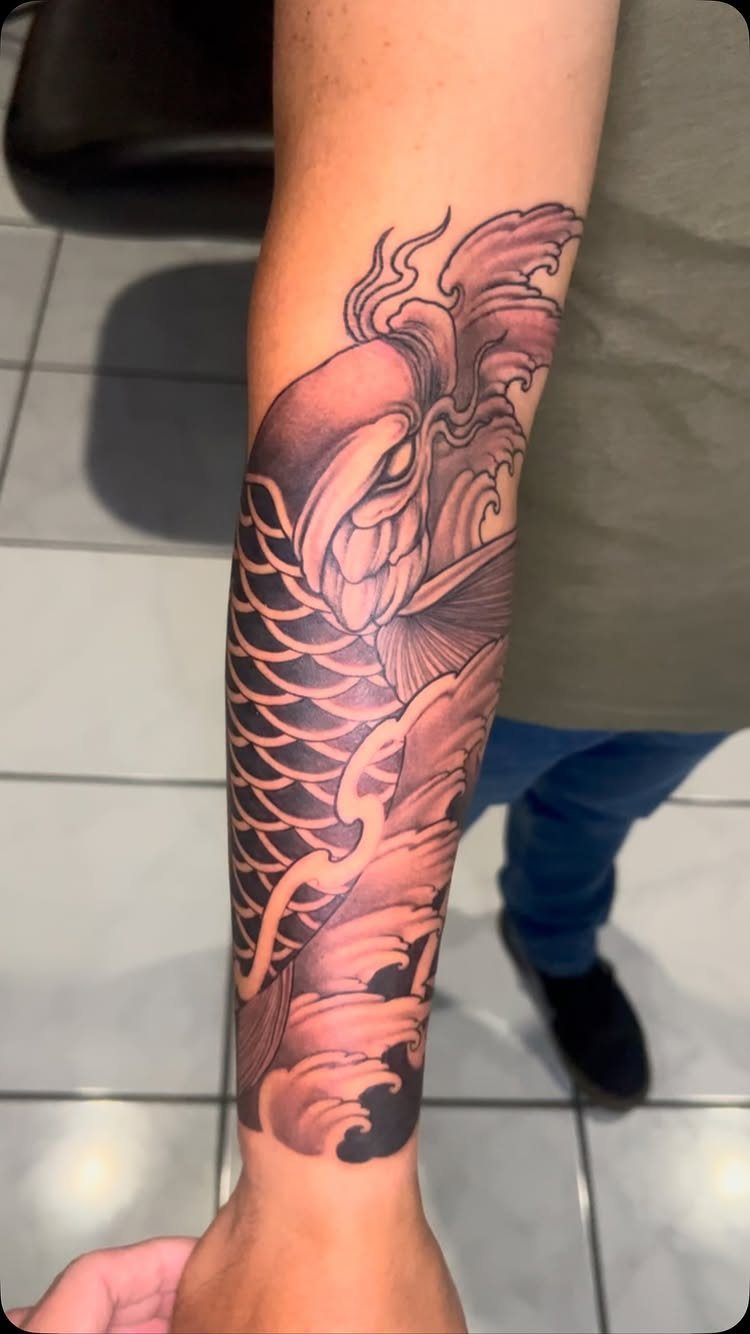 Japanese Tattoo Style: 14 Iconic Motifs and Where They Go 3 A detailed black and grey koi fish tattoo with waves on a person's forearm.