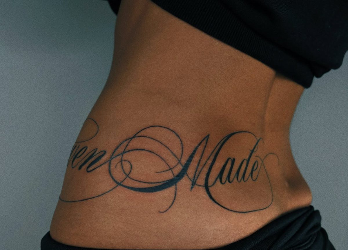 11 Lower Back Script Tattoos for Bold Ink Inspiration 10 A person's lower back displaying a large black cursive script tattoo with decorative swirls.