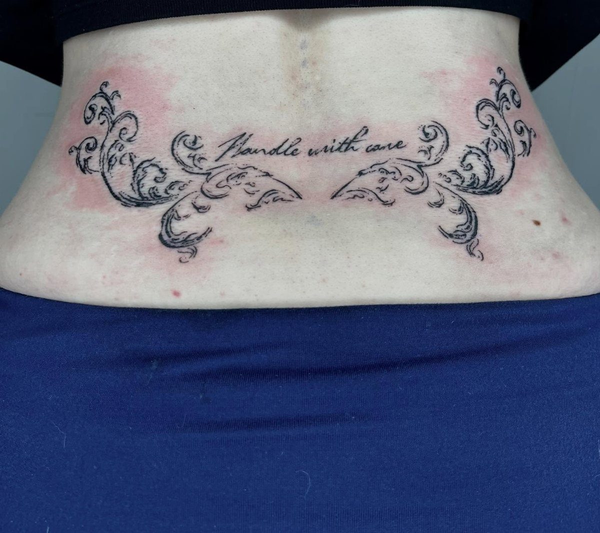 A script tattoo reading 'Handle with care' surrounded by decorative black ink swirls on a lower back.