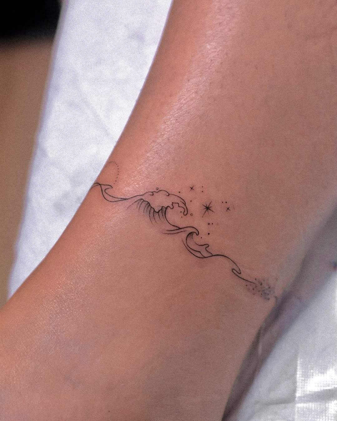 16 Front Ankle Tattoo Ideas for Women 2026 10 A minimalist black ink wave tattoo with small stars on a person's skin.