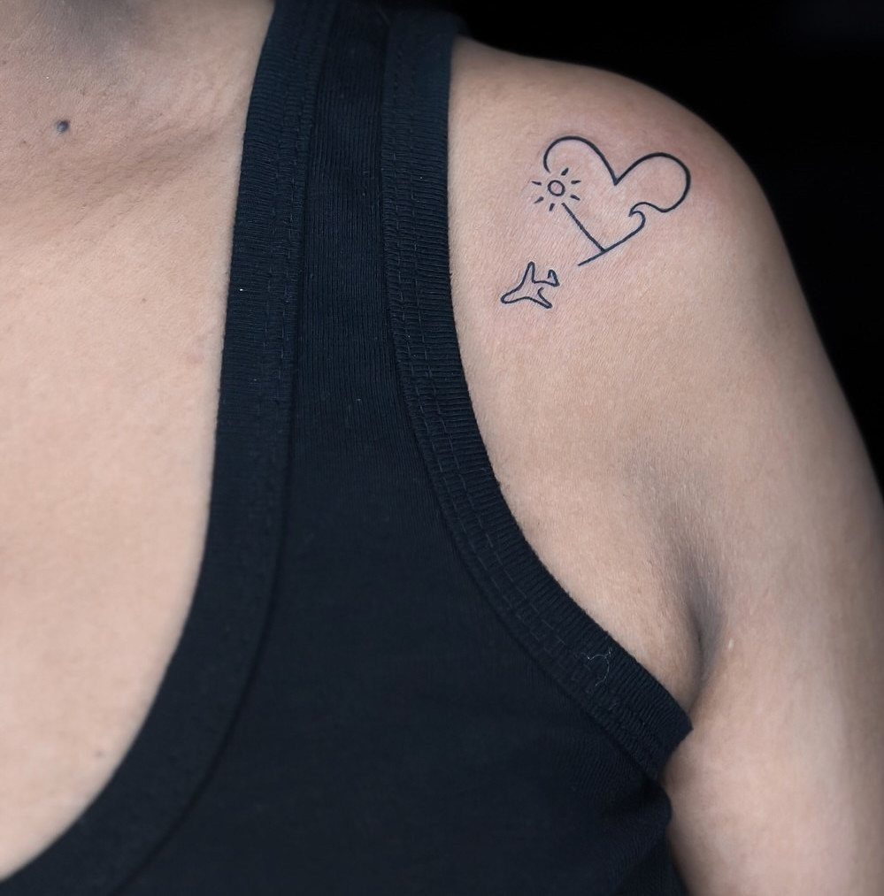 A minimalist black ink tattoo of a heart, sun, and airplane on a person's shoulder.