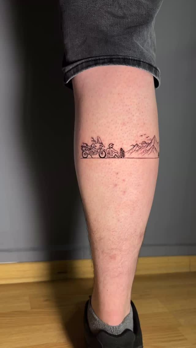 Minimalist black ink tattoo on a leg featuring a motorcycle, a person, and mountains.