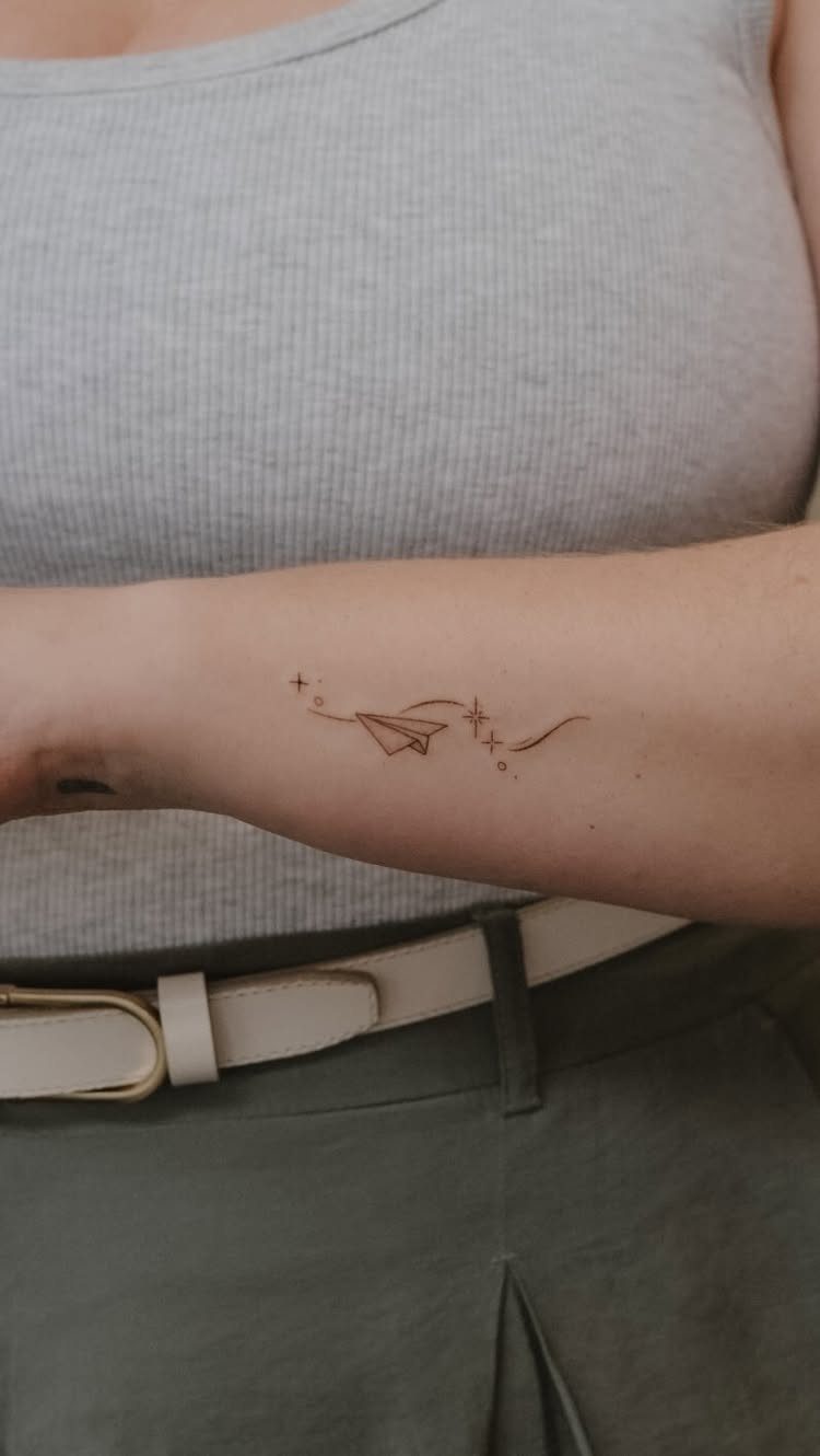 A minimalist paper plane tattoo with a dotted flight path on a person's forearm.