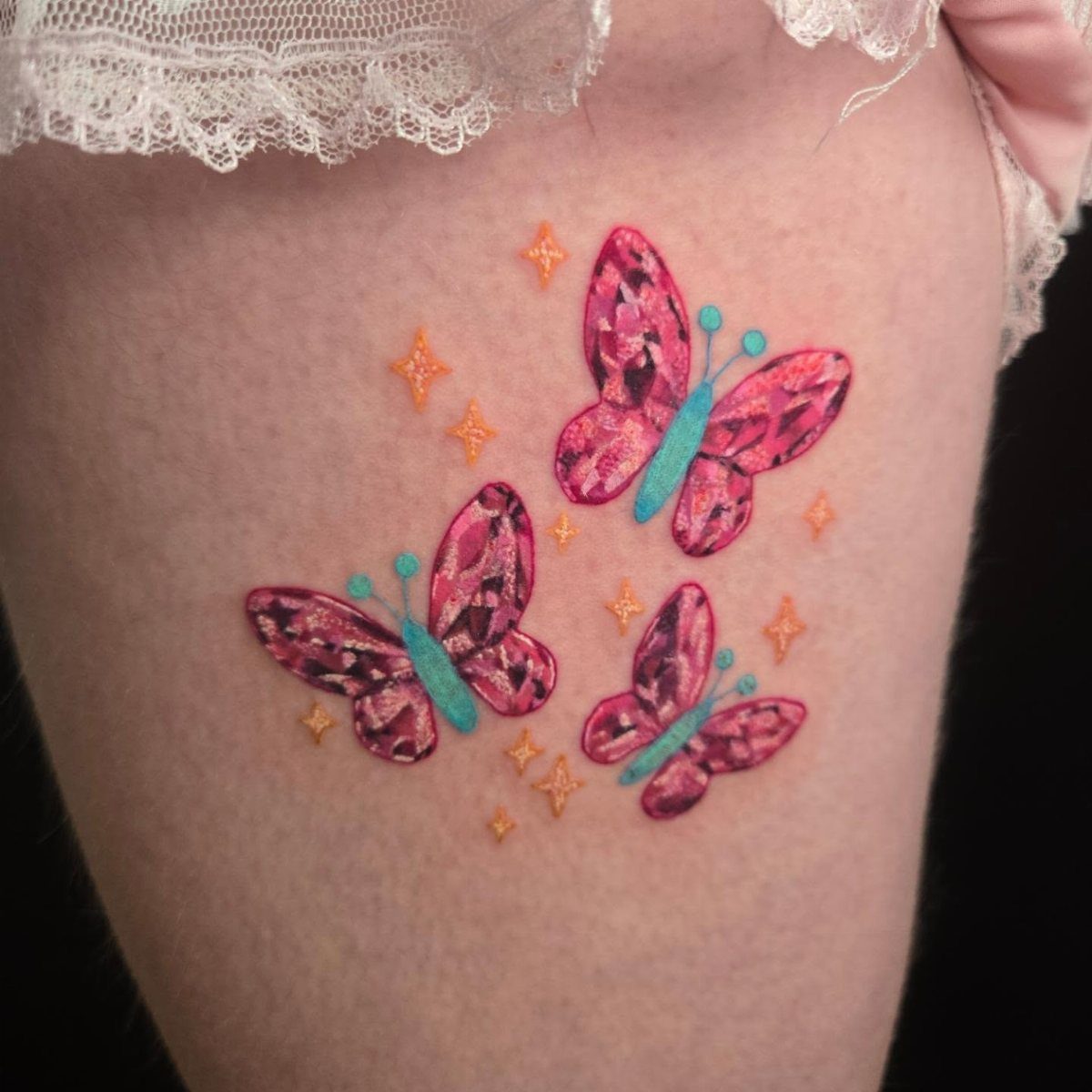 Three pink patterned butterflies with blue details and small orange stars tattooed on skin.
