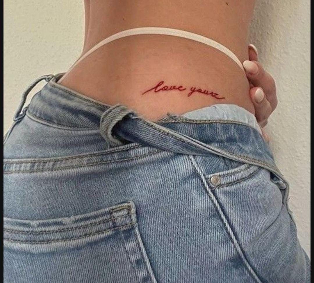 11 Lower Back Script Tattoos for Bold Ink Inspiration 9 Red cursive script tattoo reading love yours on a person's lower back above blue jeans.