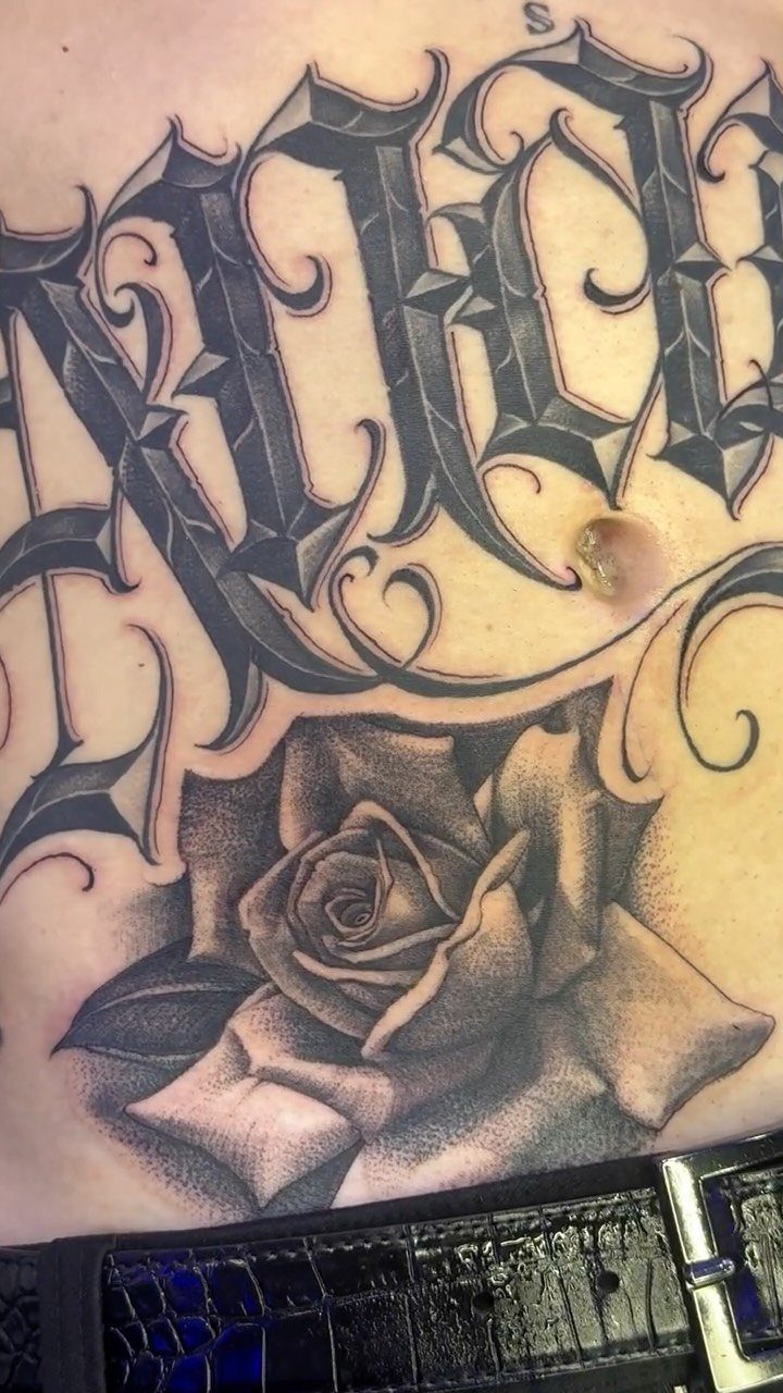 A large black and grey rose tattoo with gothic lettering on a person's stomach.