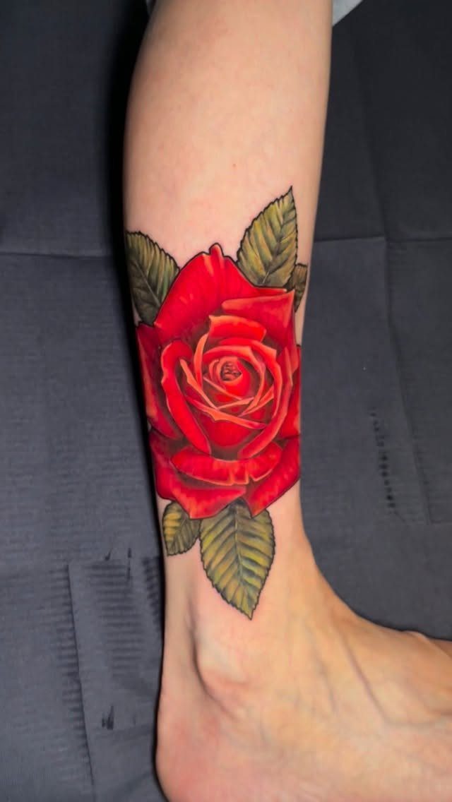 16 Front Ankle Tattoo Ideas for Women 2026 2 A large red rose tattoo with green leaves on a person's lower leg and ankle.