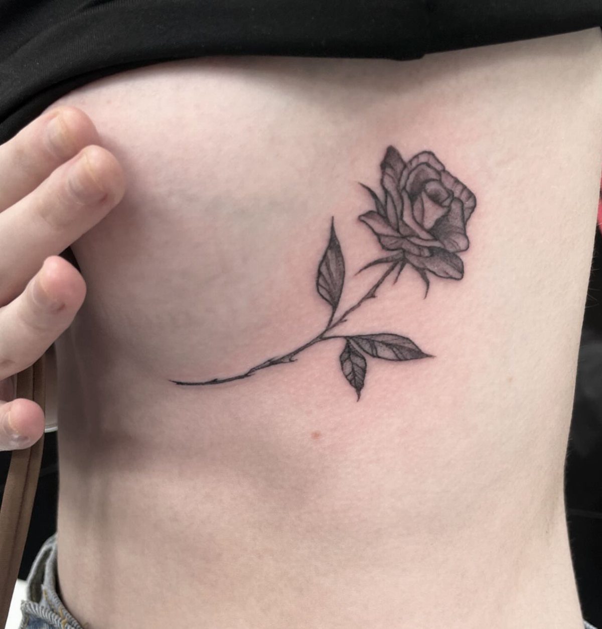 A black and grey rose tattoo with a long stem on a person's side ribs.