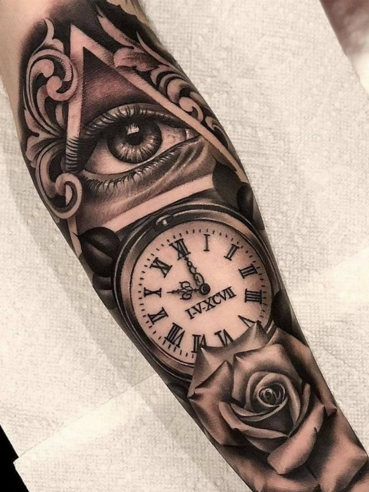 A detailed black and grey tattoo on an arm featuring an eye, a clock, and a rose.