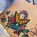 A colorful butterfly and rainbow tattoo surrounded by flowers on a person's stomach.