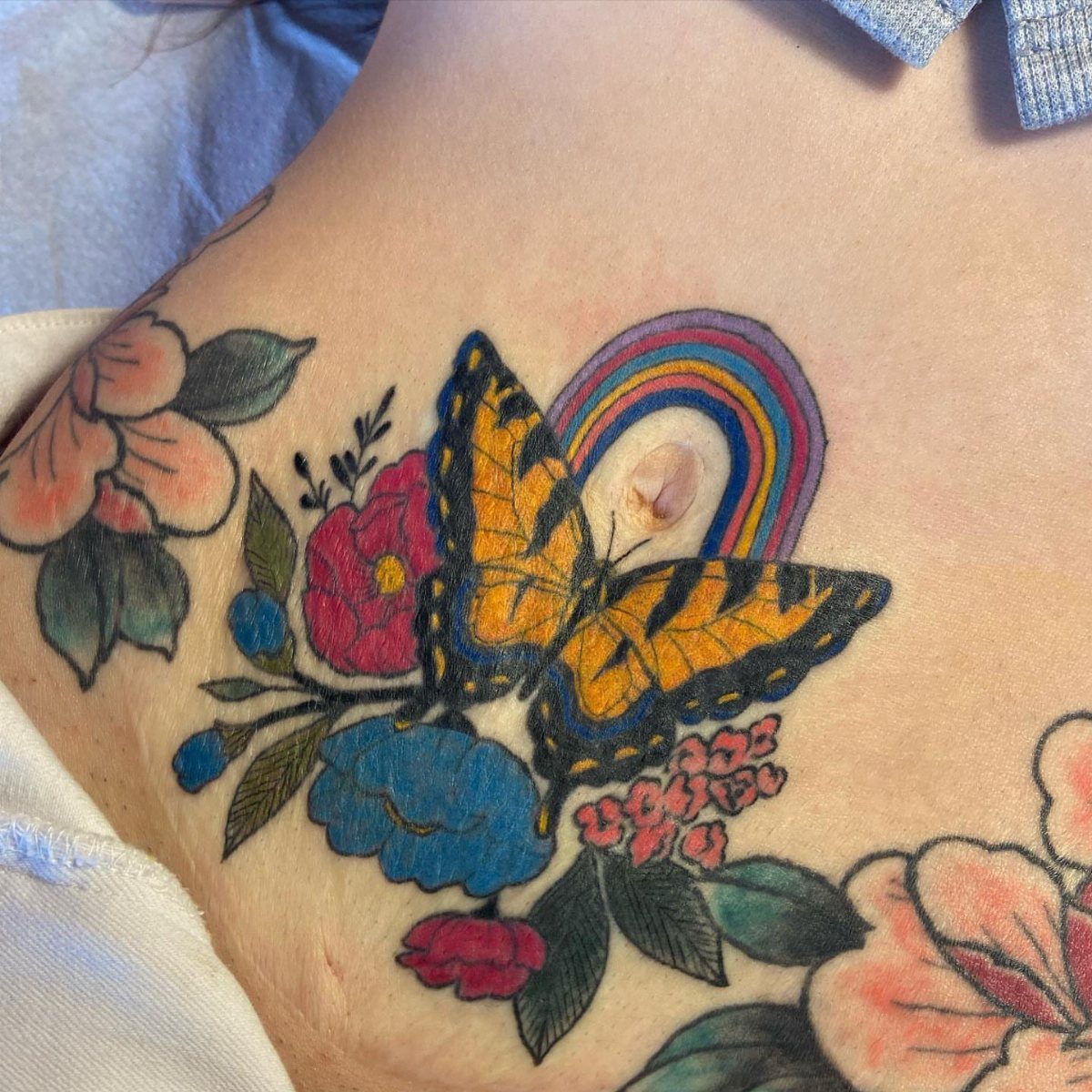 A colorful butterfly and rainbow tattoo surrounded by flowers on a person's stomach.