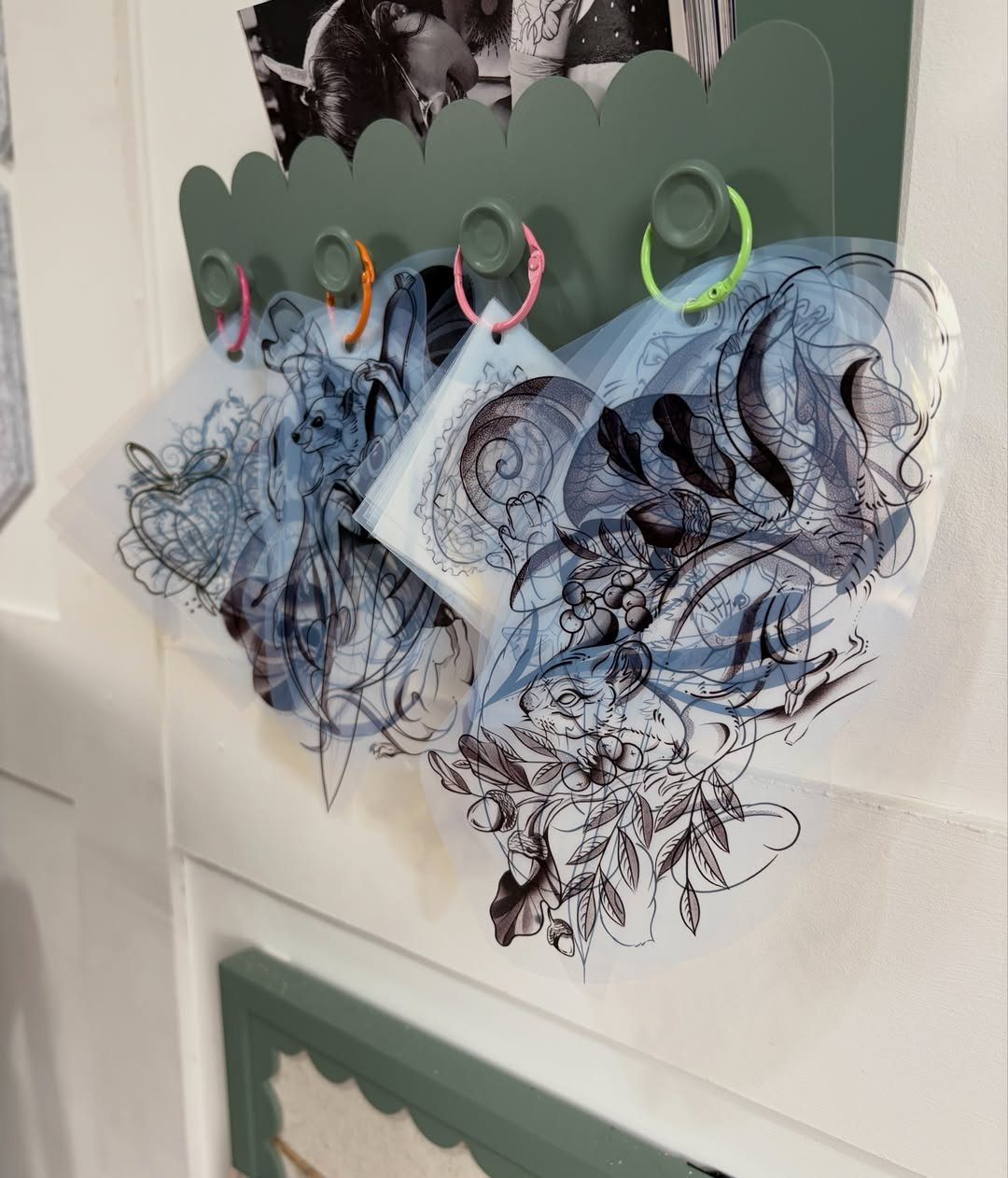 Body Tattoo Design: 5 Styles to Study Before You Book 6 Translucent tattoo stencil sheets with black ink floral designs hanging from colorful rings on a wall.