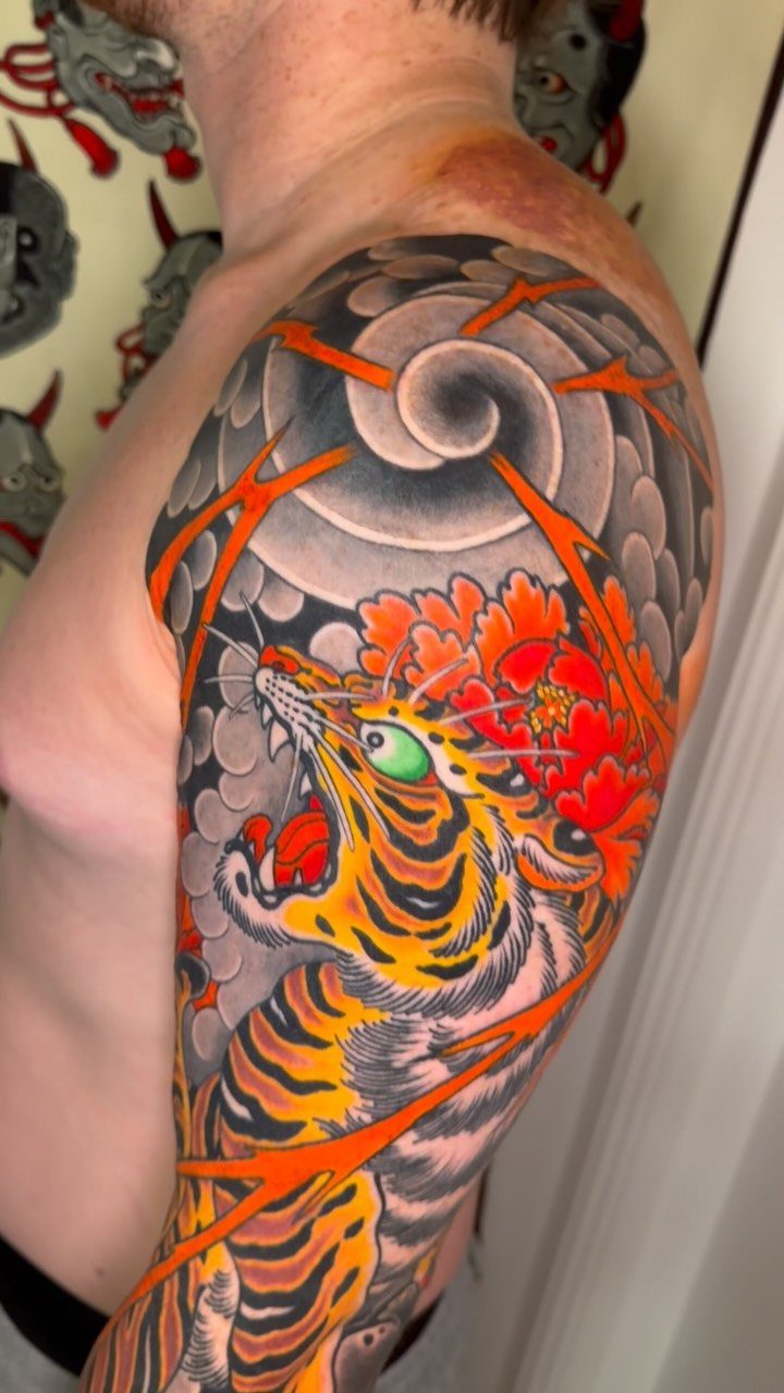 Japanese Tattoo Style: 14 Iconic Motifs and Where They Go 8 A vibrant Japanese style tiger tattoo with red peonies and grey clouds on a man's shoulder.